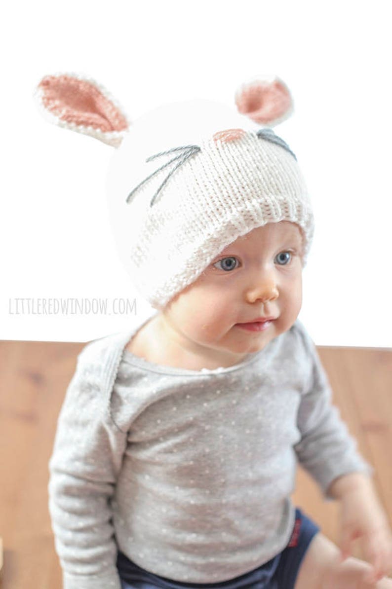 baby in gray shirt wearing a white knit bunny hat with a pink nose gray whiskers and pink lined bunny ears on top looking down at their hands  looking up and off to the right