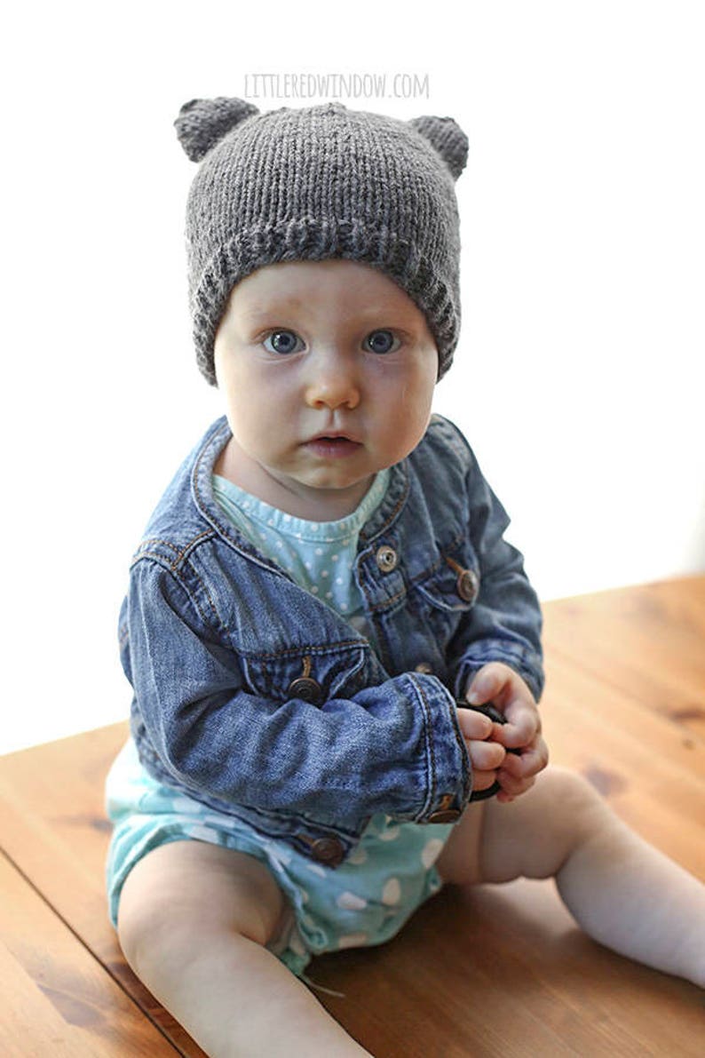 Baby looking at the camera surprised holding a lens cap and wearing a knit hat with bear ears on a wood table in front of a white background