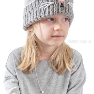 May include: A gray knitted hat with a cat face design. The hat has white whiskers, black eyes, and a pink nose.