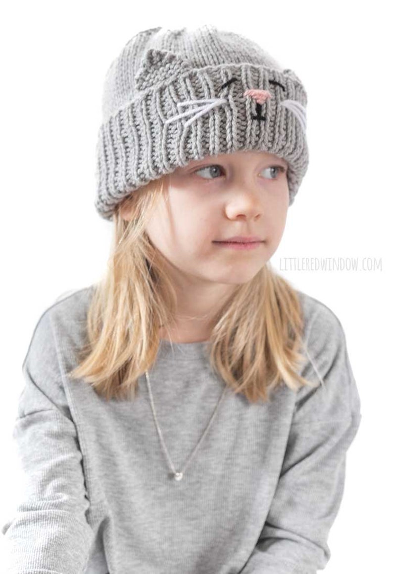 May include: A gray knitted hat with a cat face design. The hat has white whiskers, black eyes, and a pink nose.
