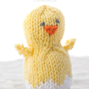 little yellow knit chick with white egg shell pattern on this bottom and two little wings on the sides sitting on a gray table