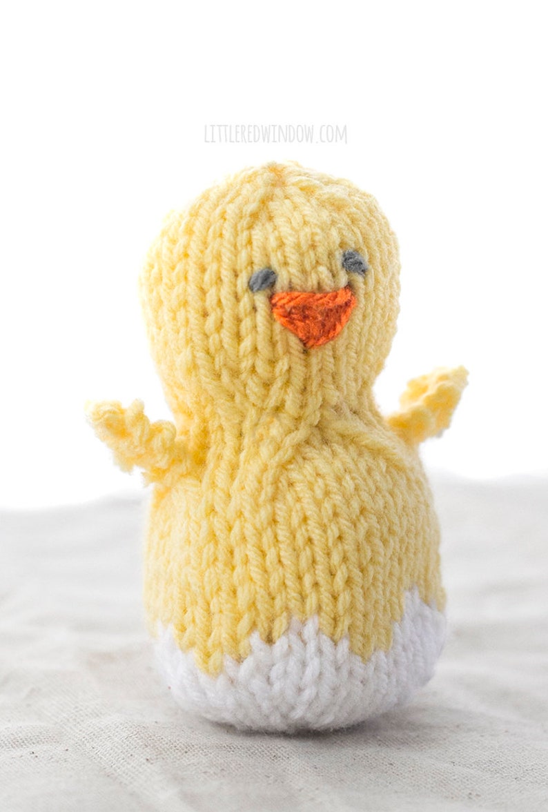 little yellow knit chick with white egg shell pattern on this bottom and two little wings on the sides sitting on a gray table