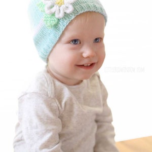 Baby in gray shirt smiling and wearing a light blue spring daisy flower hat with white band and white knit flower in front of a white background