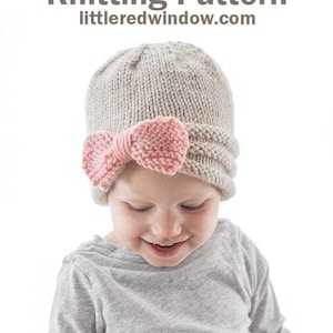 smiling girl in gray shirt wearing tan knit hat gathered in the front with large pink knit bow looking down at her lap