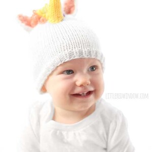 grinning baby in white shirt wearing a white knit hat with golden unicorn horn and rainbow knit mane sitting in front of a white background and looking slightly to the right