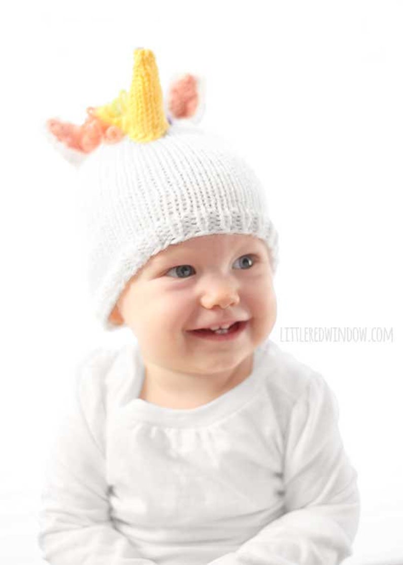 grinning baby in white shirt wearing a white knit hat with golden unicorn horn and rainbow knit mane sitting in front of a white background and looking slightly to the right