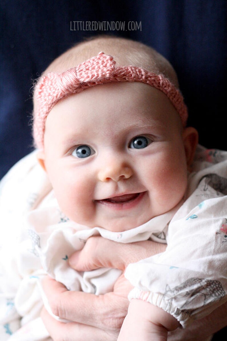 smiling chubby baby with wide blue eyes wearing a light pink ribbed knit headhand with a knit bow on the front being held in front of a dark navy blue background