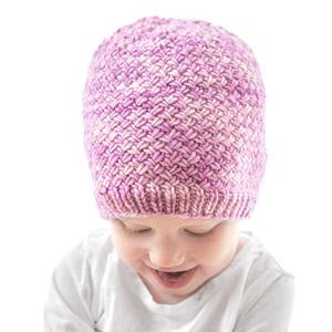 May include: A pink and white knitted baby hat with a basketweave design. The hat is worn by a child in a white long-sleeved top. Text at the top reads "Tiny Basketweave Hat Knitting Pattern" and "littleredwindow.com".