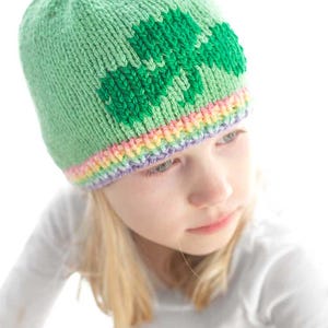 A young girl dons a light green knitted hat featuring a charming shamrock design. The hat, adorned with a rainbow band at the edge, showcases a delightful knitting pattern. Sporting a white long-sleeved shirt, she gazes slightly to the side.