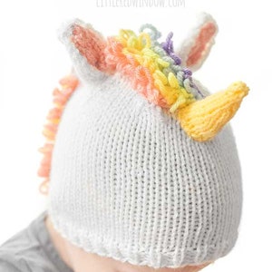 View of toddler in gray shirt wearing knit unicorn hat from slightly above on a baby showing rainbow loop stitch mane, pink and white unicorn ears and yellow knit unicorn horn