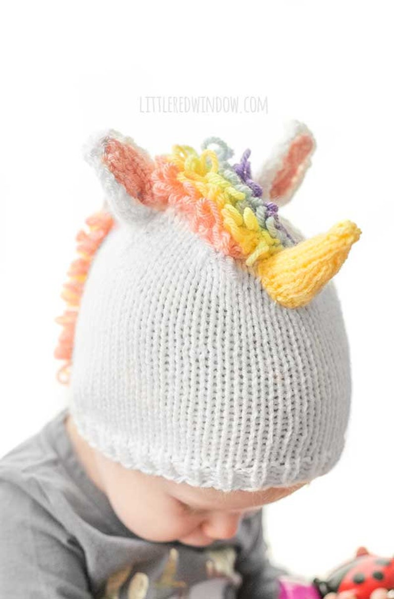 View of toddler in gray shirt wearing knit unicorn hat from slightly above on a baby showing rainbow loop stitch mane, pink and white unicorn ears and yellow knit unicorn horn