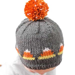 A baby wearing a knitted, dark gray candy corn hat decorated with an orange and white pattern resembling the festive treat. The top of the hat is adorned with a fluffy orange pom-pom. The background is plain white.