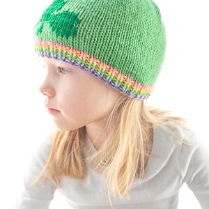 A young child with blonde hair wears a charming shamrock hat featuring a leaf design and a multicolored rainbow brim, looking to the side against a plain white background. She is dressed in a light gray long-sleeve shirt, embracing both style and comfort.