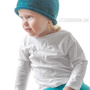 toddler in white shirt and blue pants wearing a knit hat with a rolled brim made of teal blue hand dyed yarn  sitting on a wood table and leaning over to the left