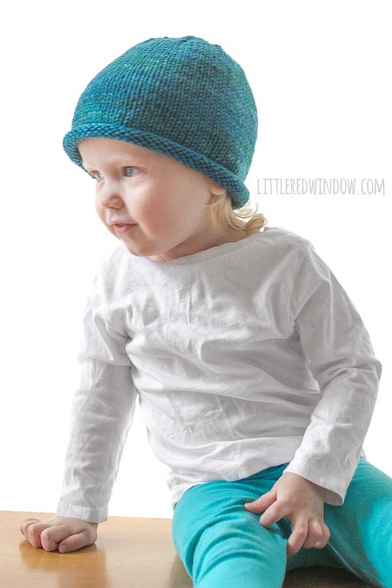 toddler in white shirt and blue pants wearing a knit hat with a rolled brim made of teal blue hand dyed yarn  sitting on a wood table and leaning over to the left
