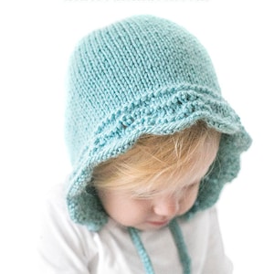 May include: A light blue knitted baby bonnet with a scalloped edge. The bonnet is worn by a child with blonde hair. The text "Bluebell Bonnet Knitting Pattern littleredwindow.com" is visible at the top of the image.