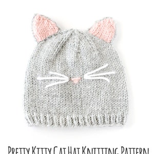 flat view of gray knit cat hat with pink ears on top and a pink nose and white whiskers on the front laying on a white background