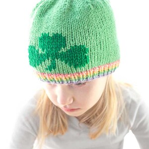 A child is sporting a green knit hat adorned with a shamrock design, showcasing a colorful rainbow  brim. The little one gazes downward, dressed in a light gray striped shirt. The vibrant, blurred background adds to this charming scene inspired by the shamrock hat knitting pattern.
