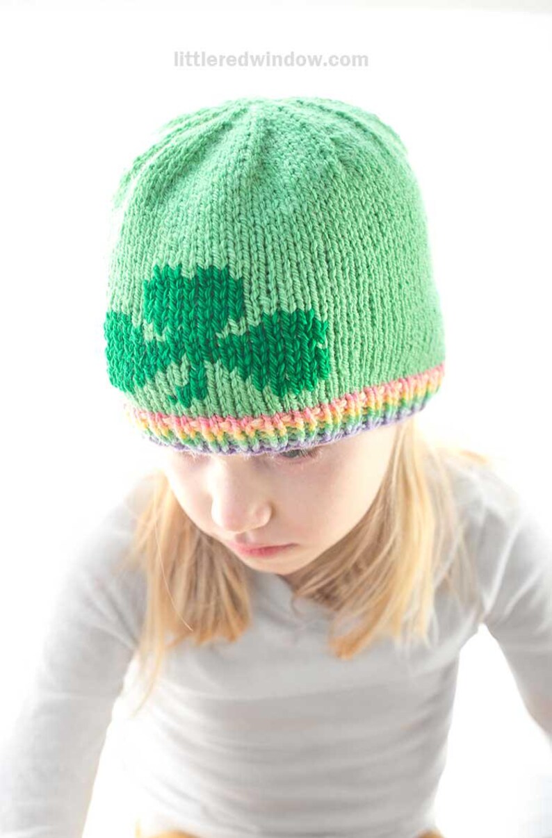 A child is sporting a green knit hat adorned with a shamrock design, showcasing a colorful rainbow  brim. The little one gazes downward, dressed in a light gray striped shirt. The vibrant, blurred background adds to this charming scene inspired by the shamrock hat knitting pattern.
