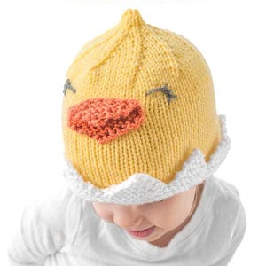 May include: A yellow knitted chick hat with a white brim. The hat has black eyes, a small orange beak, and a white brim with a scalloped edge. The hat is being worn by a child.