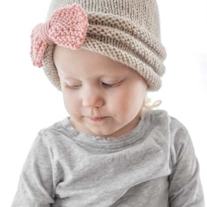 chubby girl in gray shirt wearing tan knit hat gathered in the front with large pink knit bow looking down to the left