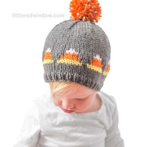 A small child wearing a candy corn hat knitted in gray with orange and white patterns and an orange pom-pom on top, looks down while dressed in a white long-sleeve shirt.