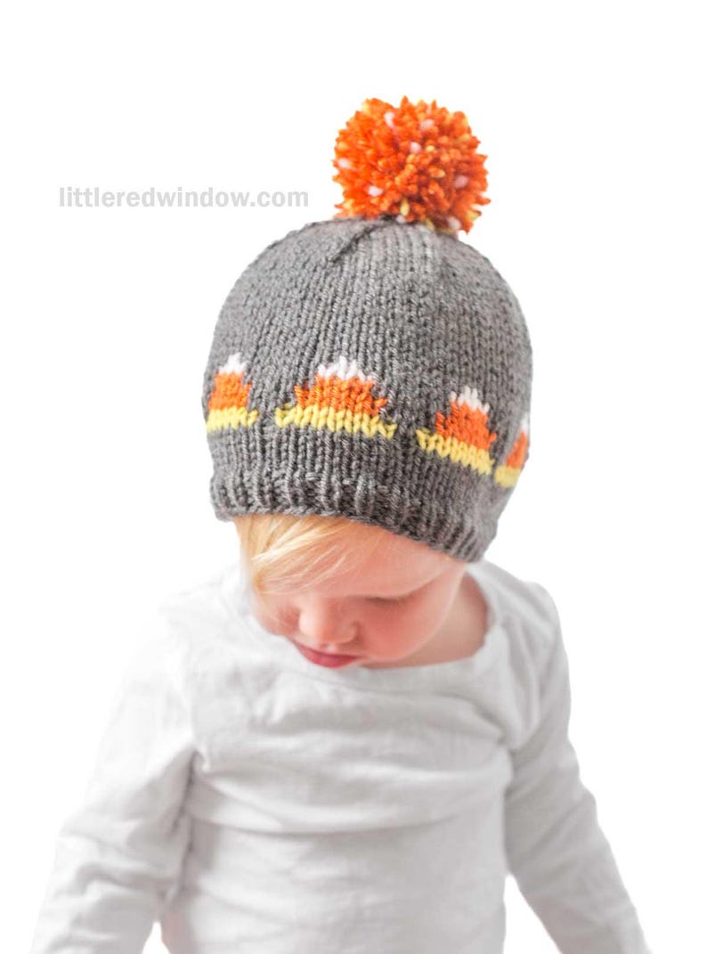 A small child wearing a candy corn hat knitted in gray with orange and white patterns and an orange pom-pom on top, looks down while dressed in a white long-sleeve shirt.
