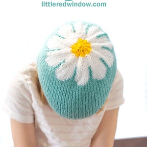 May include: A knitted hat with a daisy design on top. The hat is teal blue with a white daisy and a yellow center. The hat is being worn by a person.