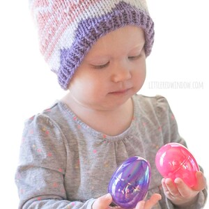 baby girl wearing knit easter egg hat and examining two pink and purple easter eggs in her hands