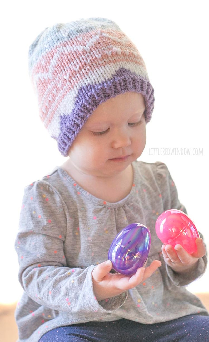 baby girl wearing knit easter egg hat and examining two pink and purple easter eggs in her hands