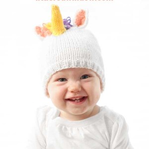 grinning baby in white shirt wearing a white knit hat with golden unicorn horn and rainbow knit mane sitting in front of a white background and looking at the camera