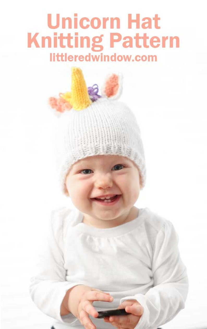 grinning baby in white shirt wearing a white knit hat with golden unicorn horn and rainbow knit mane sitting in front of a white background and looking at the camera