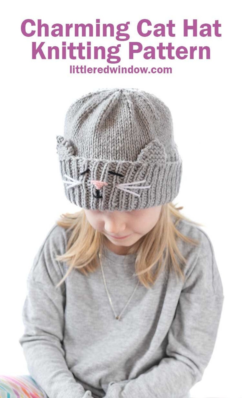 May include: A gray knitted cat hat with white whiskers and a pink nose. The hat is being worn by a child with blonde hair.