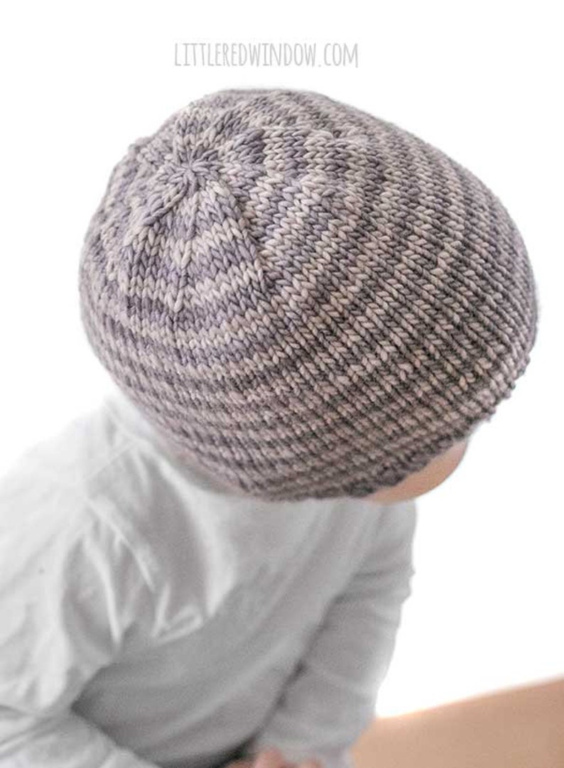 top view of toddler in white shirt wearing a tan and medium brown thin striped knit hat of a white background