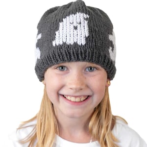 May include: A grey knitted hat with white ghost designs. The hat is worn by a person. The text "Gentle Ghosts Hat Knitting Pattern" and "littleredwindow.com" are displayed at the top of the image.
