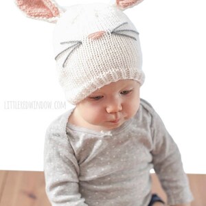 baby in gray shirt wearing a white knit bunny hat with a pink nose gray whiskers and pink lined bunny ears on top looking down at their hands  looking down