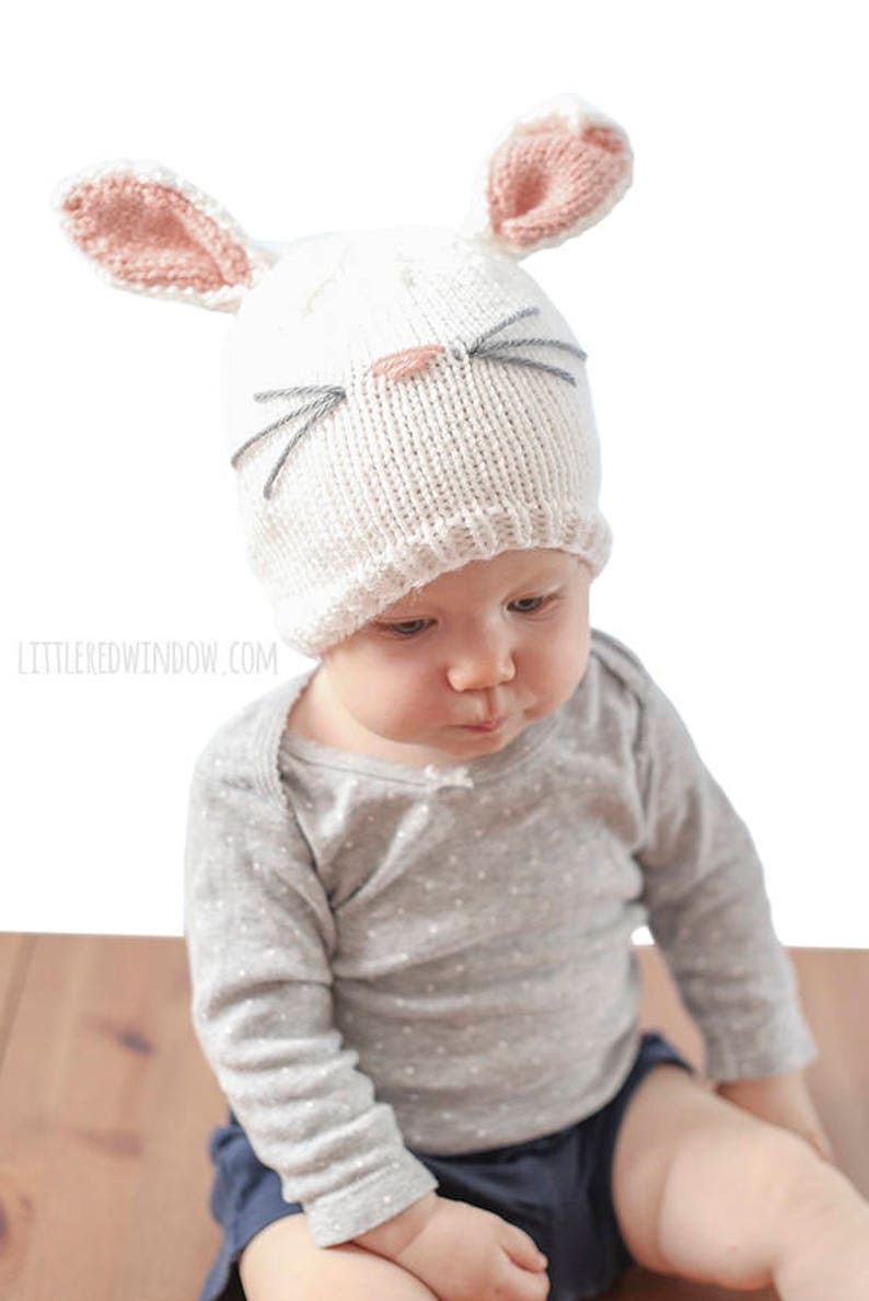 baby in gray shirt wearing a white knit bunny hat with a pink nose gray whiskers and pink lined bunny ears on top looking down at their hands  looking down