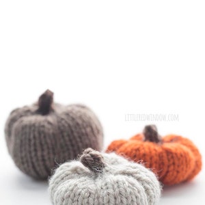 three small knit pumpkins knit with orange tan and medium brown yarn with dark brown knit pumpkin stems sitting on a white table with the tan pumpkin in front
