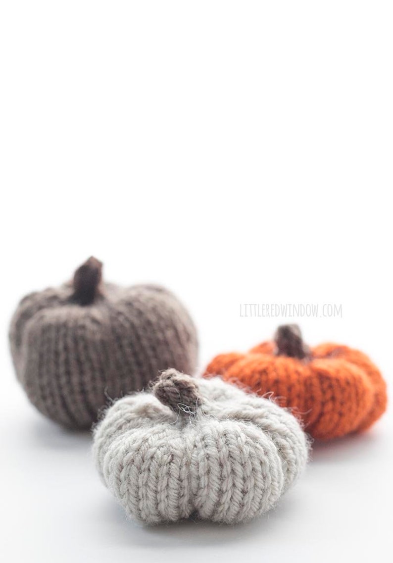 three small knit pumpkins knit with orange tan and medium brown yarn with dark brown knit pumpkin stems sitting on a white table with the tan pumpkin in front