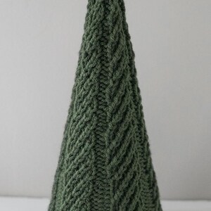 The large Cozy Christmas tree knitting pattern features a super easy twisted rib pattern in a tree shape knit in dark green yarn sitting on a white table in front of a gray wall