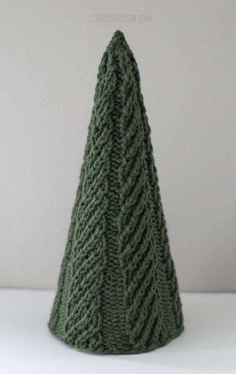 The large Cozy Christmas tree knitting pattern features a super easy twisted rib pattern in a tree shape knit in dark green yarn sitting on a white table in front of a gray wall