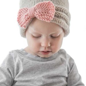 smiling girl in gray shirt wearing tan knit hat gathered in the front with large pink knit bow looking down to the right