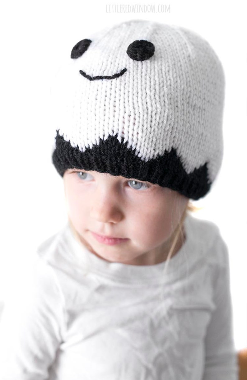 K&ouml;nnte beinhalten: Ein Kind tr&auml;gt eine wei&szlig;e und schwarze gestrickte Geisterm&uuml;tze mit schwarzen Augen und einem l&auml;chelnden Mund.