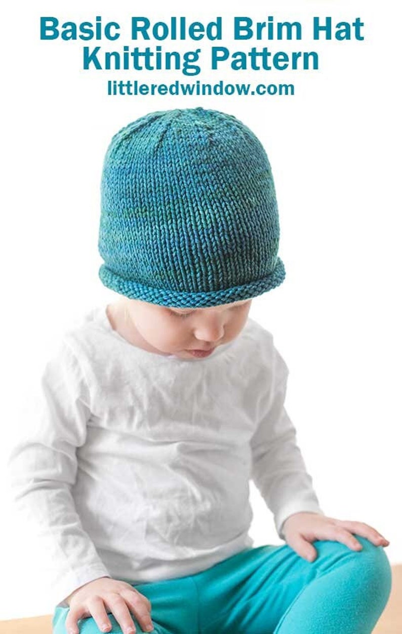 toddler in white shirt and blue pants wearing a knit hat with a rolled brim made of teal blue hand dyed yarn  sitting on a wood table and looking down at their lap