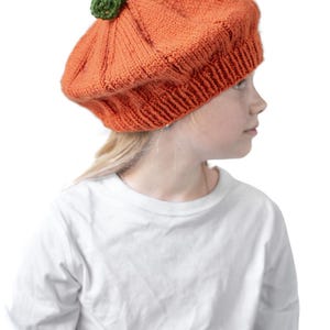 Può includere: Un berretto lavorato a maglia di colore arancione a forma di zucca con un bordo a coste e un piccolo stelo verde sulla parte superiore. Il berretto presenta creste verticali e un motivo a maglia testurizzato. Il testo littleredwindow.com è visibile.
