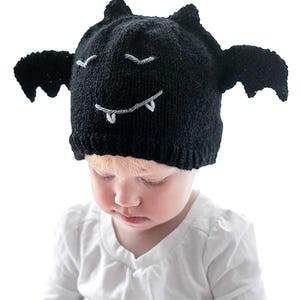 A young child is wearing a black bat hat designed to look like a bat, complete with wings and a face. The child is looking down, wearing a white long-sleeve shirt, and holding an item in their hands. The background is plain white.