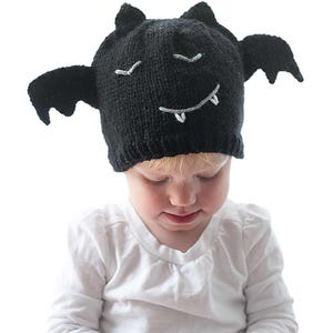 A young toddler wearing a charming black knit bat hat with wings, white eyes, and a smiling mouth with fangs holds an object while looking down.