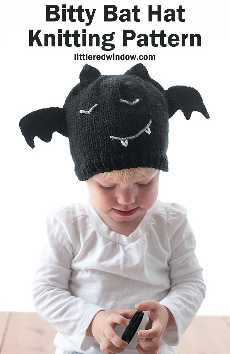 A young toddler wearing a charming black knit bat hat with wings, white eyes, and a smiling mouth with fangs holds an object while looking down.