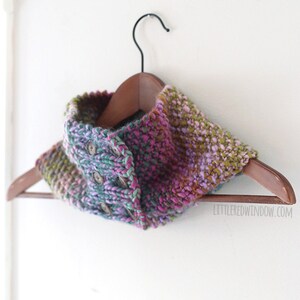 May include: A multi-colored knitted cowl with buttons hanging on a wooden hanger. The cowl is made with a chunky yarn and has a gradient of colors from green to purple to pink.
