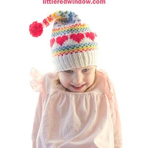 May include: A child wearing a white knit hat with a rainbow stripe pattern and pink hearts. The hat has a pom-pom on top. The hat is titled "Rainbow Love Stocking Cap Knitting Pattern" and the website "littleredwindow.com" is displayed below the title.